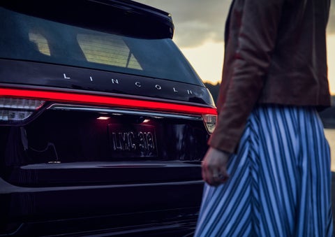 A person is shown near the rear of a 2024 Lincoln Aviator® SUV as the Lincoln Embrace illuminates the rear lights | Crossroads Lincoln of Southern Pines in Southern Pines NC